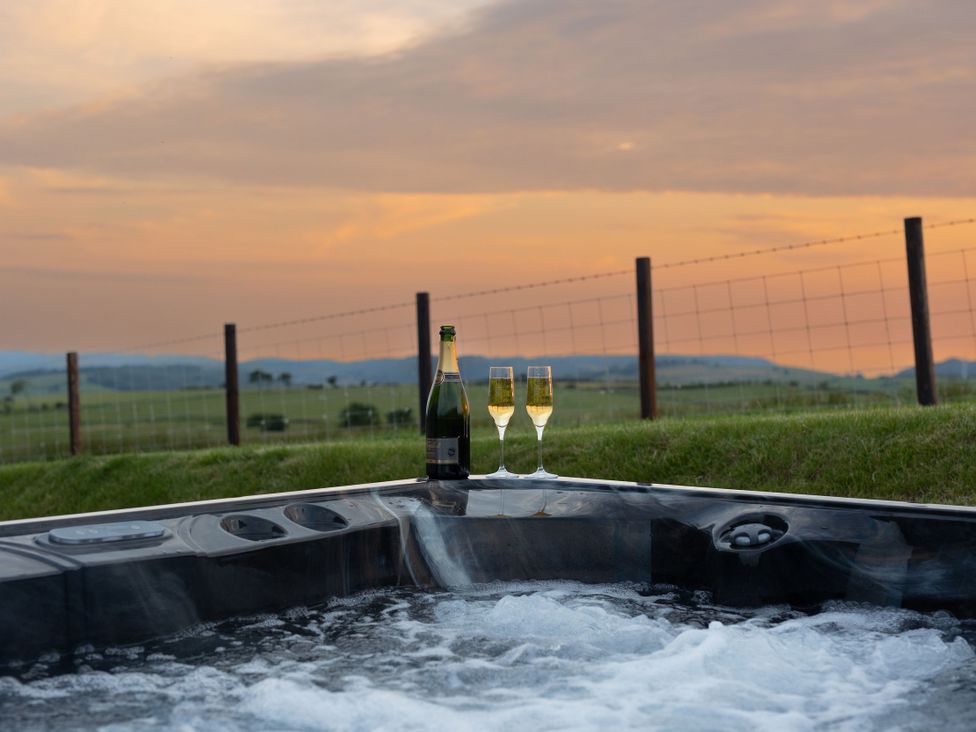 A hot tub with champagne glasses and a bottle against a sunset landscape at Golygfa Cwrt Talybont
