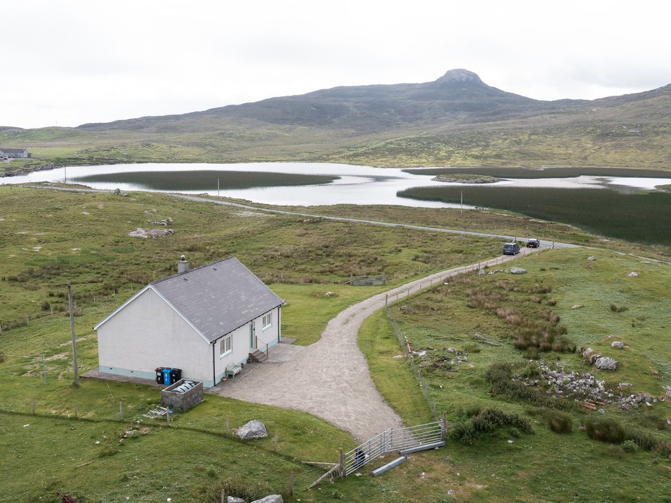 A house near a lake with a dirt road and cars at Taigh Eva near Lochmaddy