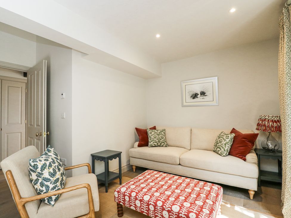 A living room with a beige sofa with cushions a wooden armchair with a cushion a patterned ottoman small tables and a framed picture on the wall at Cedar in Elcot near Newbury