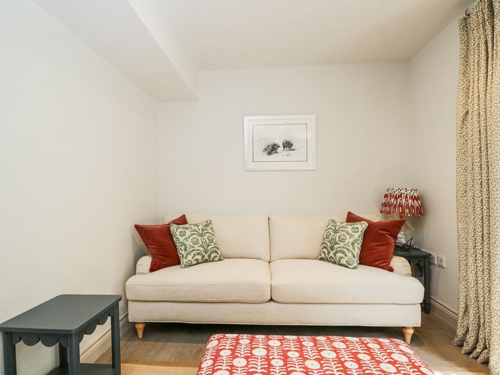 A beige sofa with green and red cushions next to a small dark side table with a lamp and curtains in the corner of a room