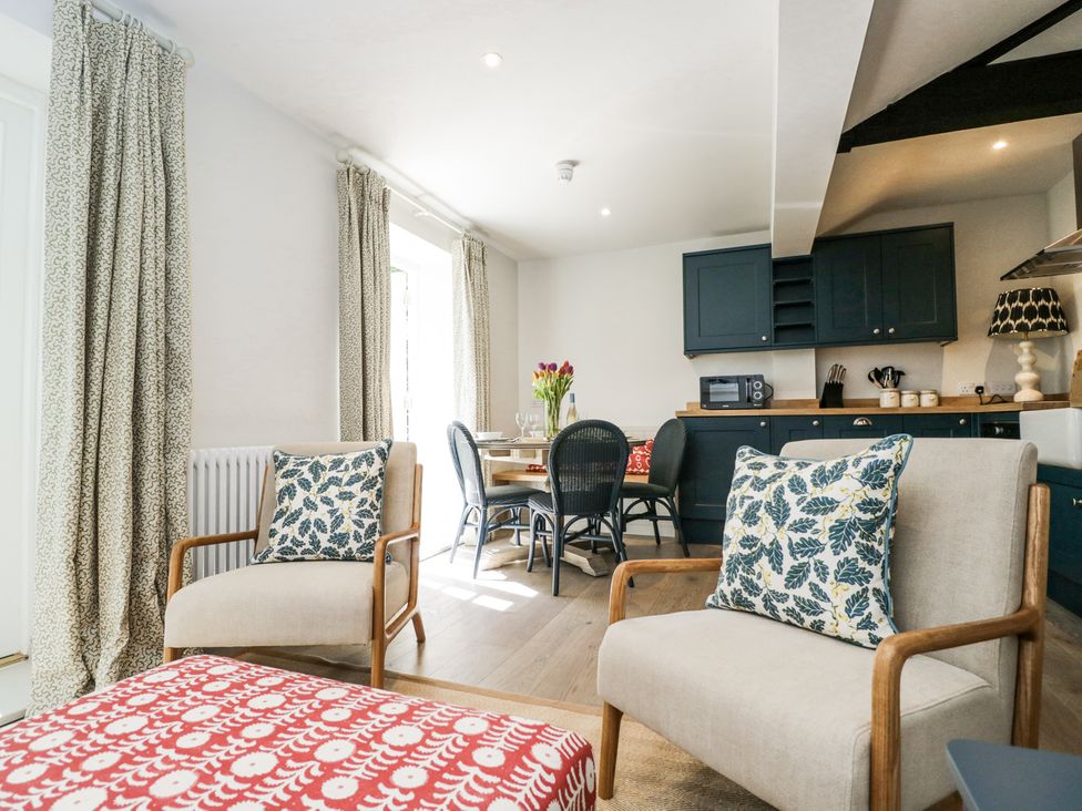 A sitting area with two armchairs with leaf-patterned cushions and a red patterned ottoman in front of a dining table and kitchen at Cedar in Elcot near Newbury