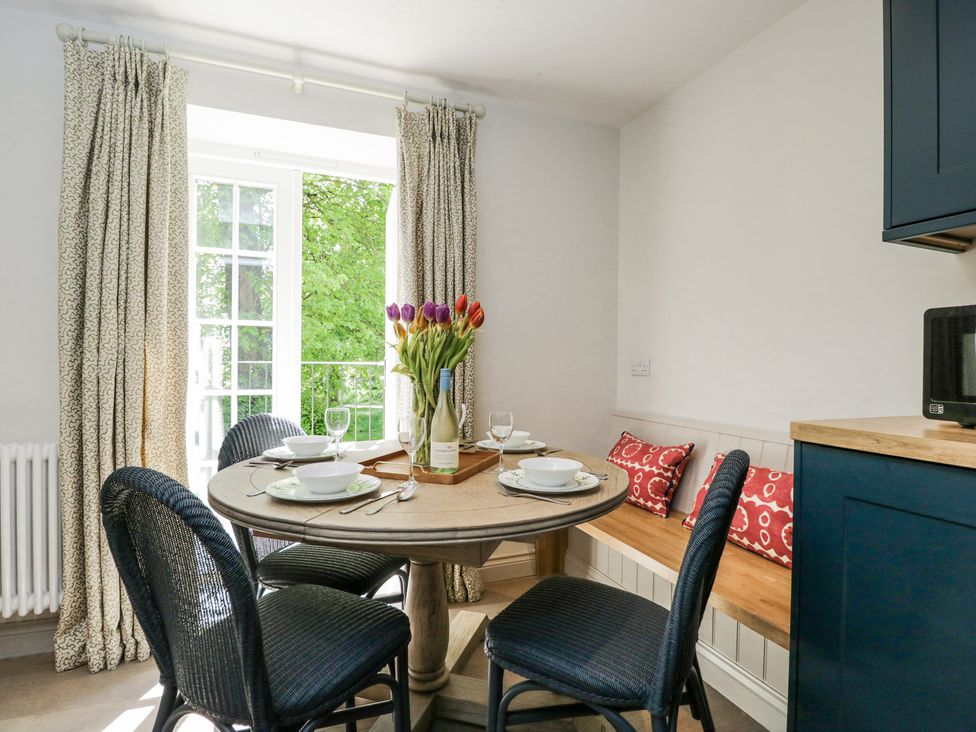 A dining area with a round wooden table set with plates and bowls four black wicker chairs a built in wooden bench with red patterned cushions a vase of multicolored tulips and a bottle of wine near a window with patterned curtains at Cedar in Elcot near Newbury