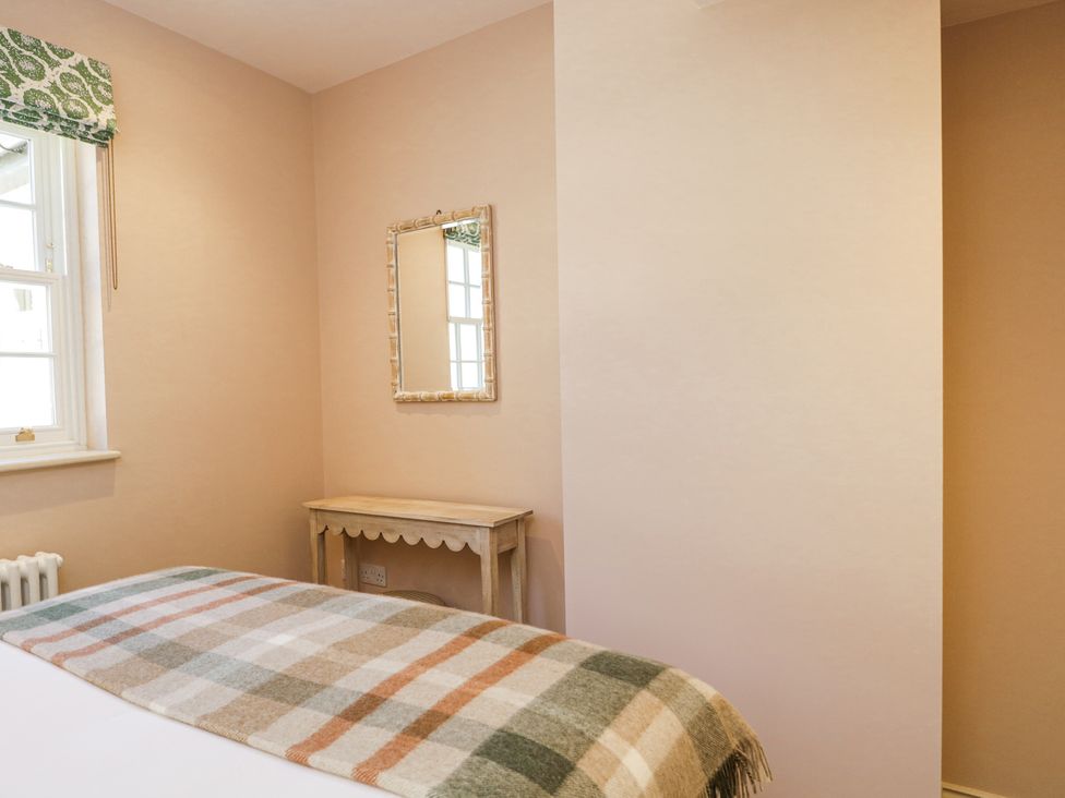 A bedroom with a bed covered by a plaid blanket a window with a patterned blind a wooden table and a framed mirror on the wall at Cedar in Elcot near Newbury
