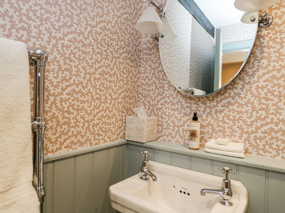 A bathroom sink with two taps a round mirror with wall lights wallpaper and a towel rail with a towel at Cedar Elcot near Newbury