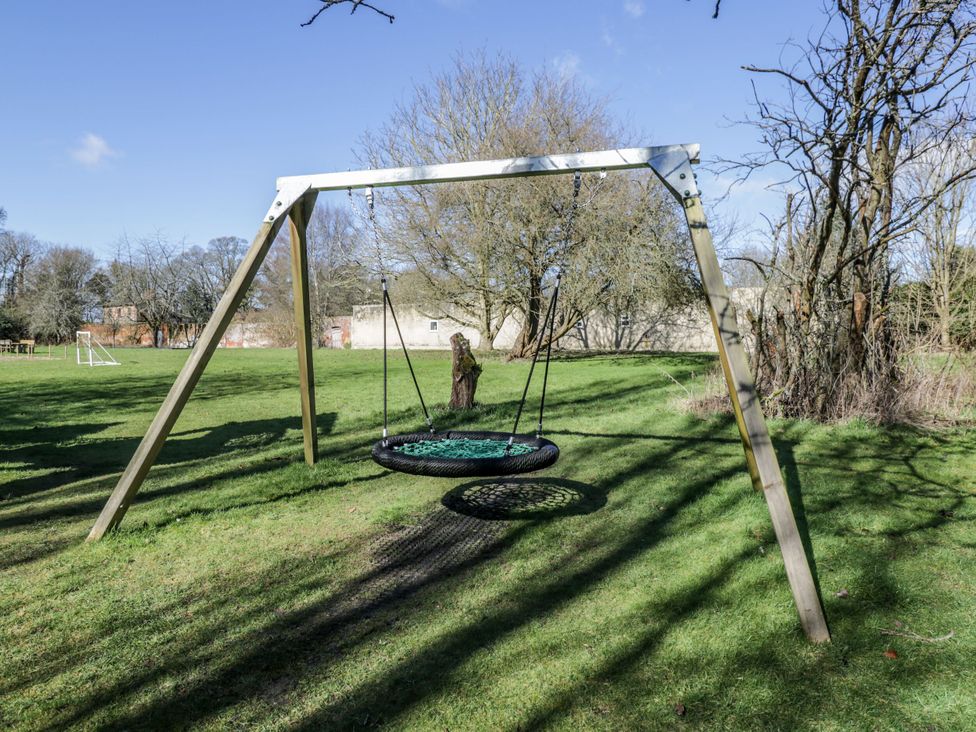 An outdoor swing set with a circular net swing on grass with trees and a building in the background at Cedar in Elcot near Newbury