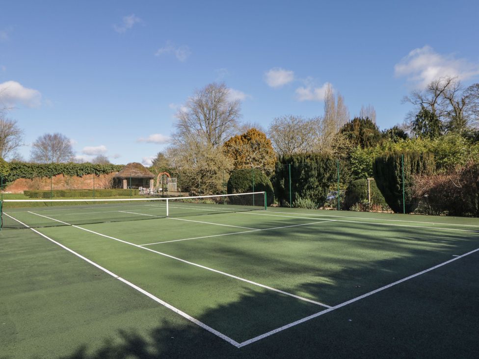 An outdoor tennis court with surrounding trees and a small thatched gazebo in the background at Cedar in Elcot near Newbury