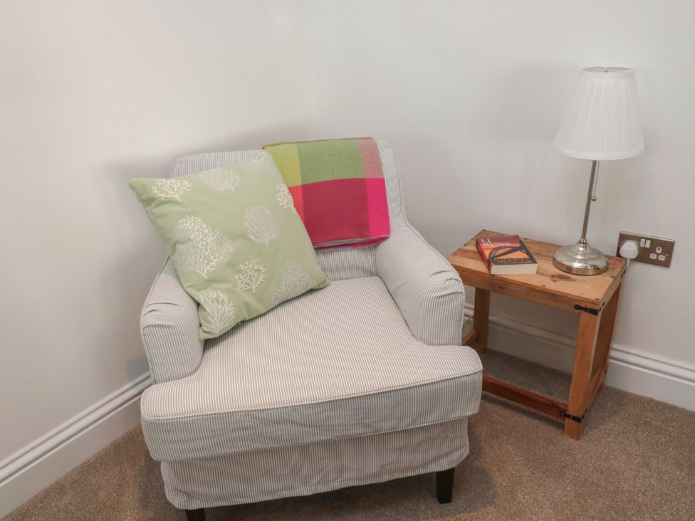 An armchair with a cushion and blanket beside a side table with a lamp and book at Harvest Cottage 2 in Berwick-upon-Tweed