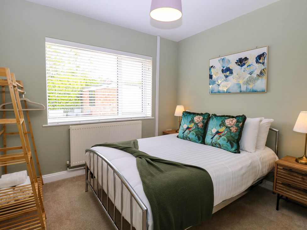 A bedroom with a bed with bird pillows and green blanket two bedside tables with lamps wooden clothes rack and window blinds at Little Lodge in Dersingham