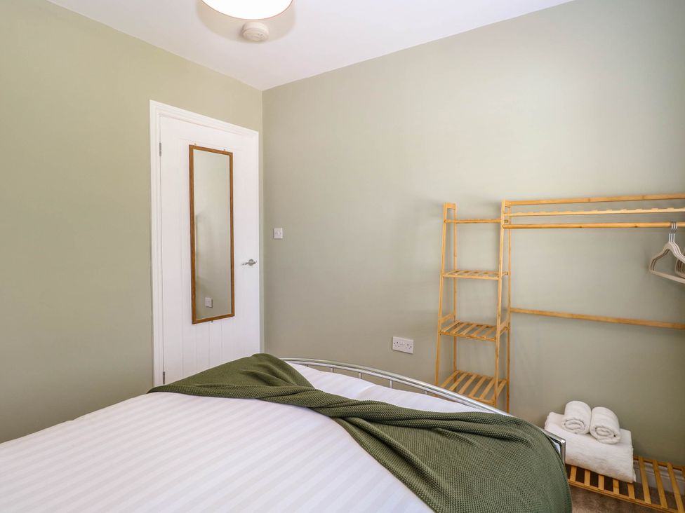 A bedroom with a bed covered with a green blanket a door with a mirror and a wooden clothes rack with hangers and folded towels at Little Lodge in Dersingham
