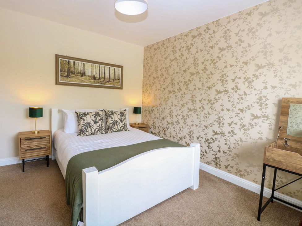 A bedroom with a white bed frame green blanket and floral cushions two wooden bedside tables with green lamps and floral patterned wallpaper at Little Lodge in Dersingham