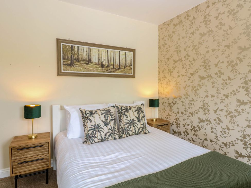 A bedroom with a bed with palm tree patterned pillows two bedside tables with lamps and a painting of a forest above the bed at Little Lodge in Dersingham