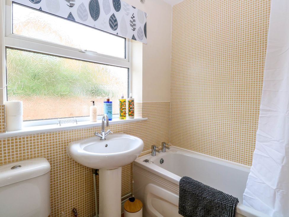 A bathroom with a pedestal sink, a bathtub with a towel hanging on the side, a toilet, and a window with frosted glass and patterned curtains at Little Lodge in Dersingham