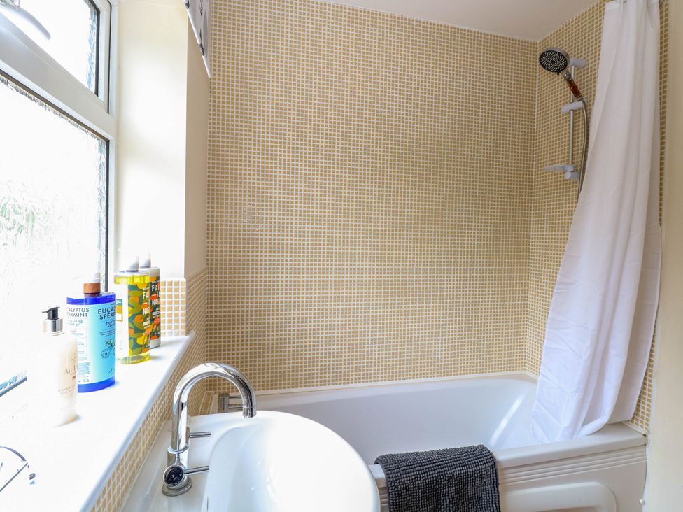 A bathroom with a bathtub shower combination with a white shower curtain and small yellow tiled walls with toiletries on the window ledge at Little Lodge in Dersingham
