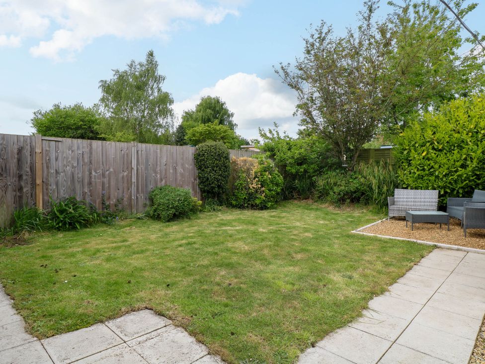 A backyard with grass lawn, a wooden fence, bushes, trees, and outdoor seating on gravel at Little Lodge in Dersingham