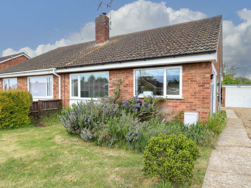 A brick bungalow house with windows and a chimney surrounded by a garden with shrubs and a lawn at Little Lodge in Dersingham