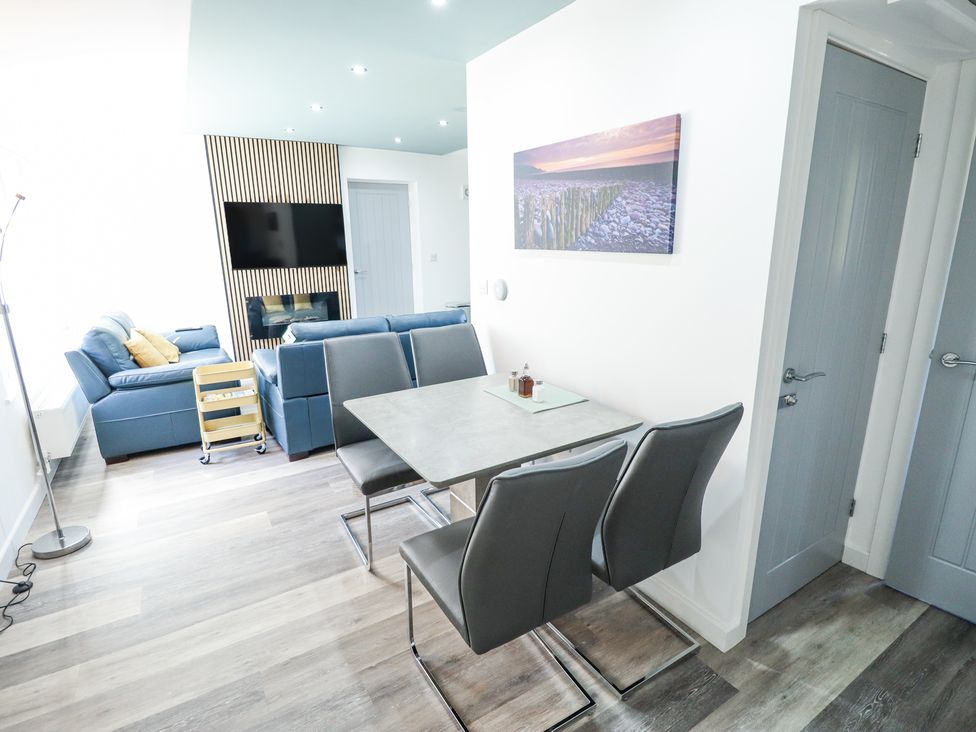 A dining area with a table and four chairs next to a living room with blue sofas and a wall-mounted TV at 19 The Court in Anderby Creek