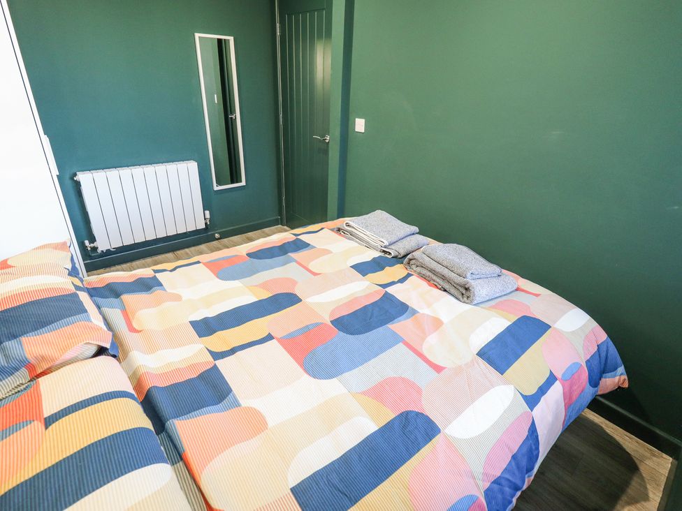 A bedroom with a bed covered in a colorful geometric patterned duvet two folded towels on the bed a green wall a door a wall-mounted radiator and a rectangular mirror at 19 The Court in Anderby Creek