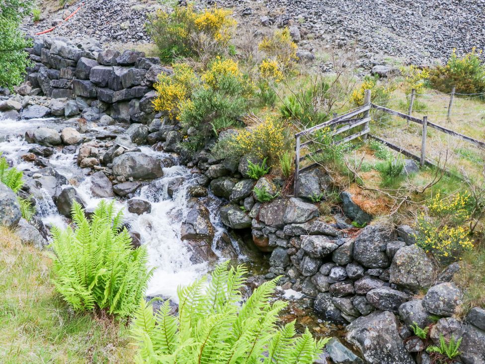 A stream flowing over rocks with ferns and a wooden fence at Swirral Edge Cottage in Glenridding