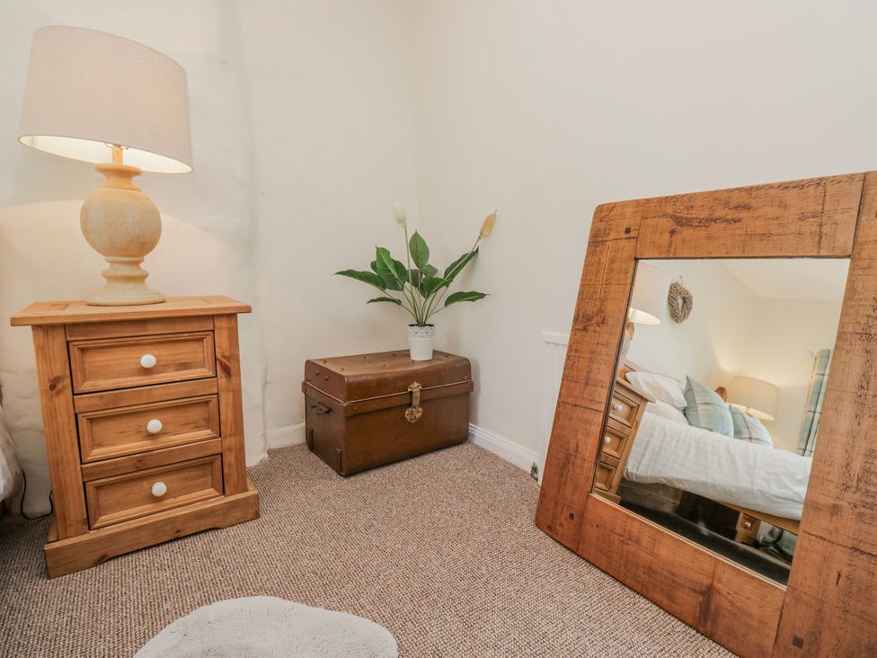 A bedroom with a lamp, side table, mirror, chest, and plant at High Ickenthwaite Farmhouse Ulverston