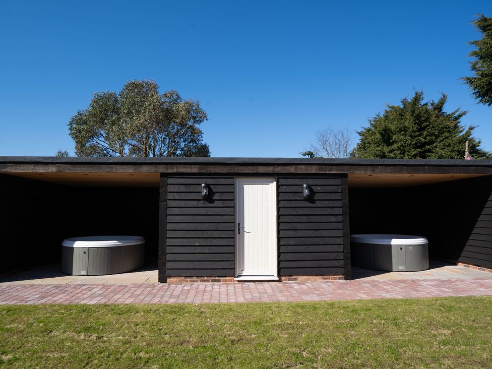 Two hot tubs under a black wooden shelter with a central white door and a brick path in front at Coral Cottage in Kessingland