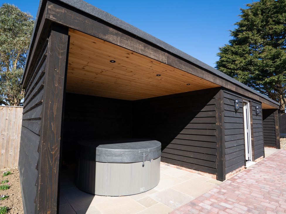 An outdoor wooden structure with a covered area housing a hot tub on a stone and brick patio at Coral Cottage in Kessingland