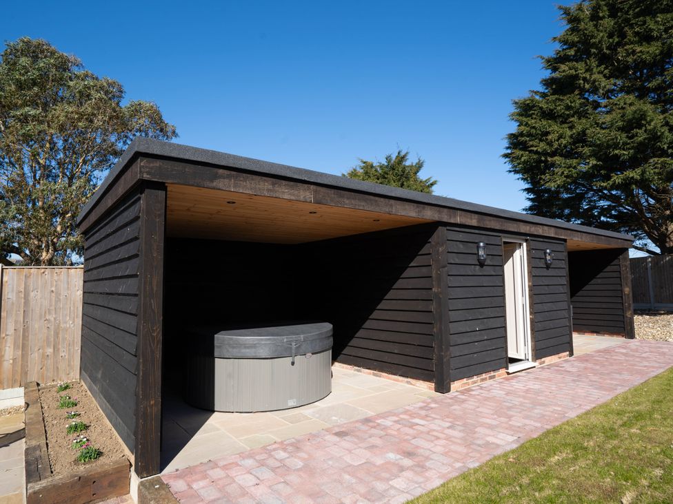 An outdoor wooden structure with a covered area housing a hot tub next to a small flower bed at Coral Cottage in Kessingland