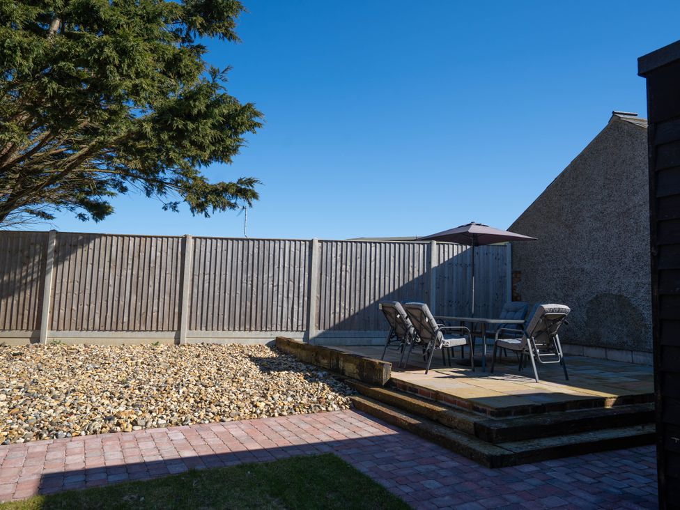 A backyard with a patio table and chairs under an umbrella on a raised stone platform and a gravel area with a wooden fence in Coral Cottage in Kessingland