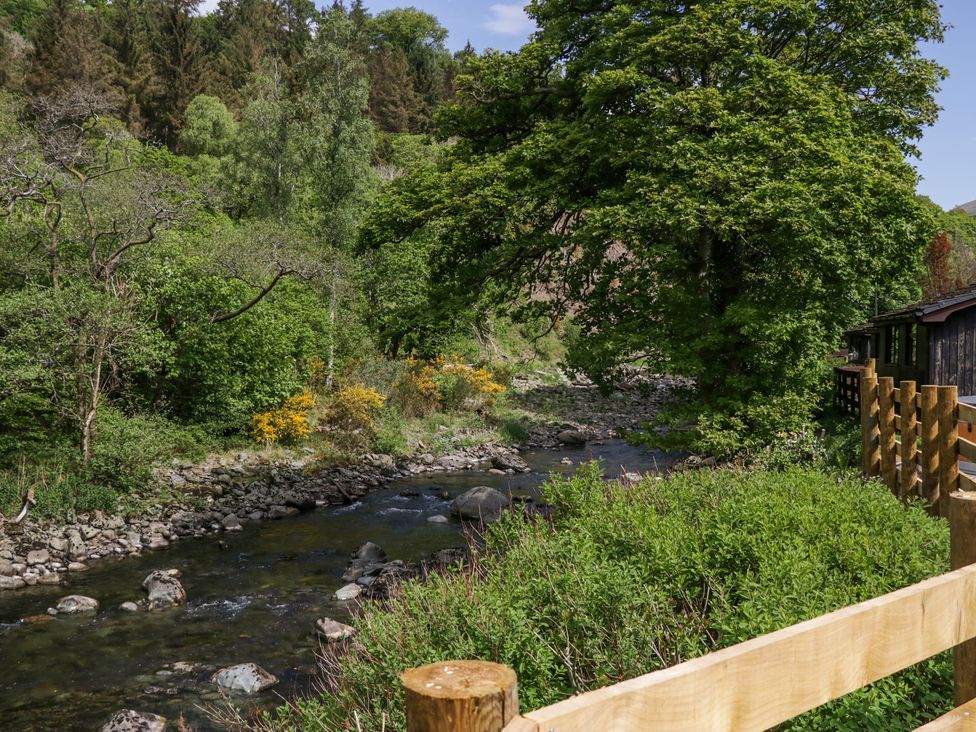 A river with rocks surrounded by trees and bushes with a wooden fence and cabin on the right at Elter Luxury Pod in Keswick