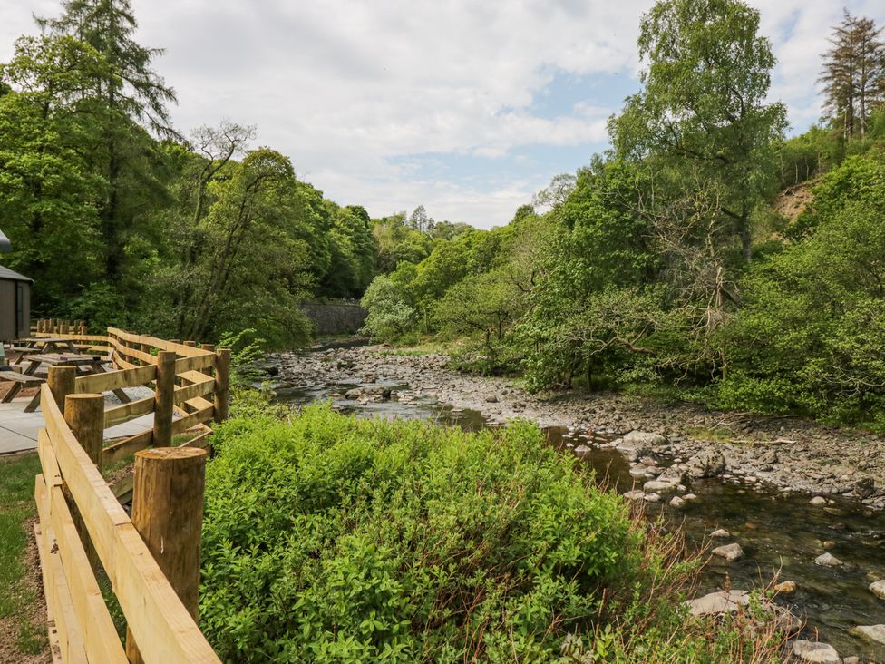 A wooden fence next to a rocky river with trees and bushes at Elter Luxury Pod in Keswick