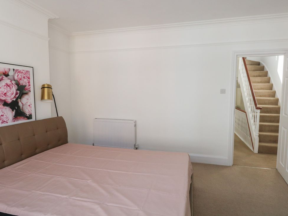 A bedroom with a bed covered with a pink sheet a framed floral picture on the wall and a staircase visible through an open door at Seaside House in Weymouth