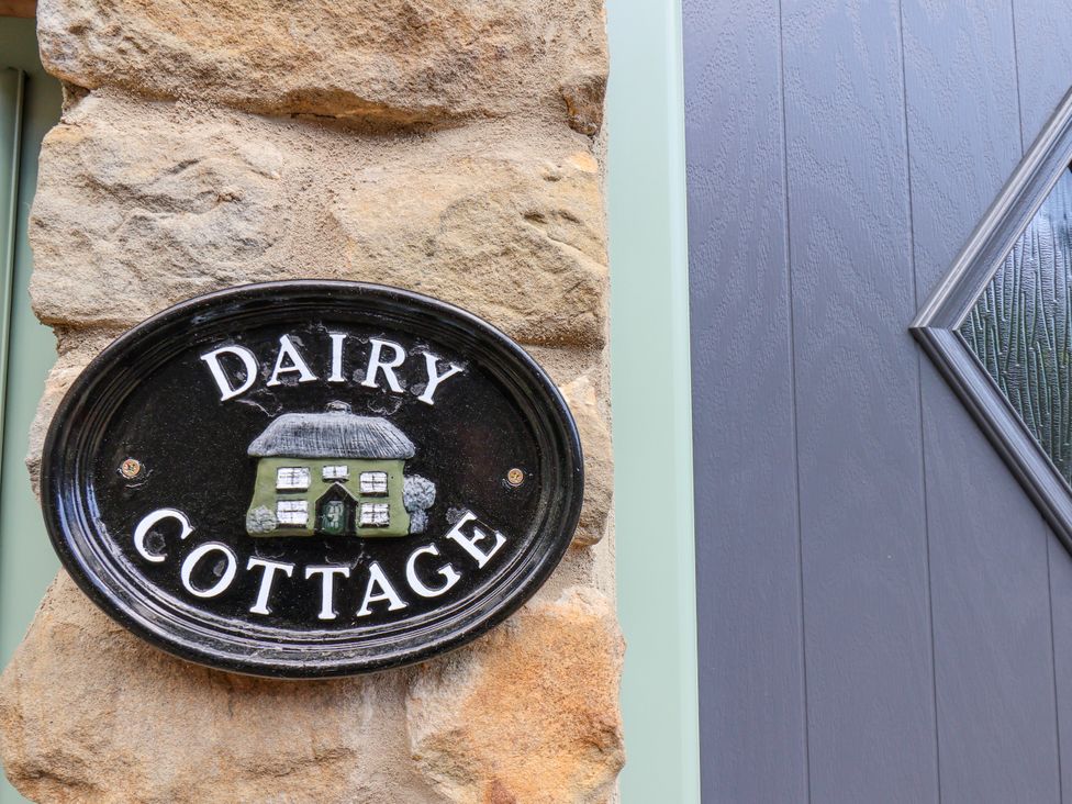 A sign reading Dairy Cottage mounted on a stone wall next to a gray door with a diamond-shaped window at Wayside Farm Dairy Cottage Cloughton near Burniston