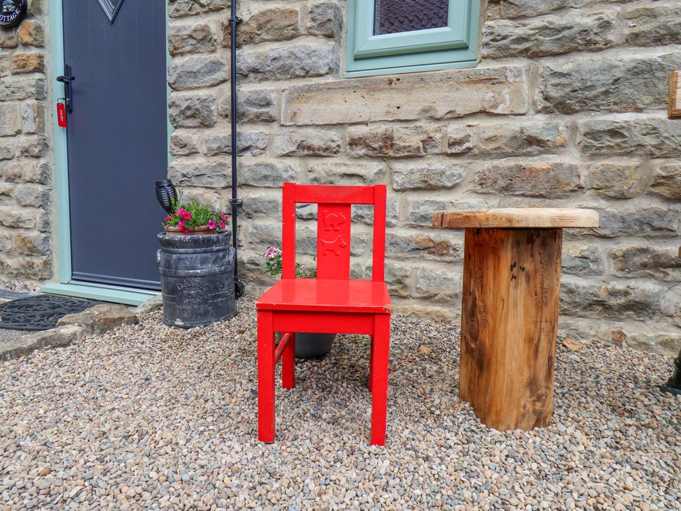 A red chair and a wooden stump table on gravel outside a stone wall with a gray door and small window at Wayside Farm Dairy Cottage in Cloughton near Burniston