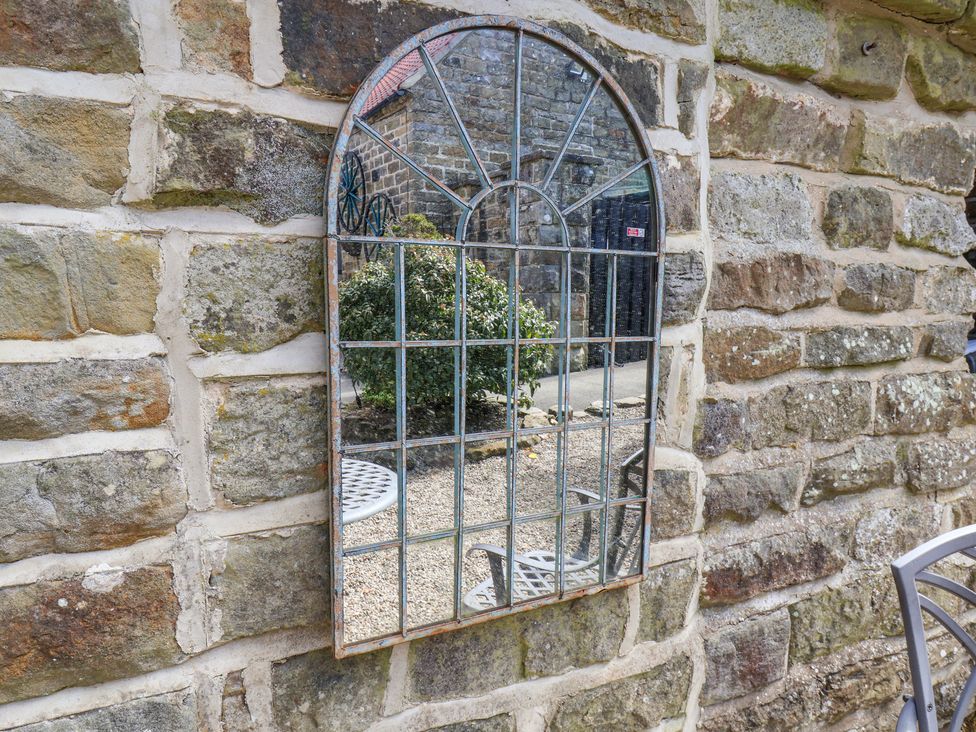 A metal framed arched mirror mounted on a stone wall reflecting outdoor furniture bushes and a stone building at Wayside Farm Stable Cottage Cloughton near Burniston
