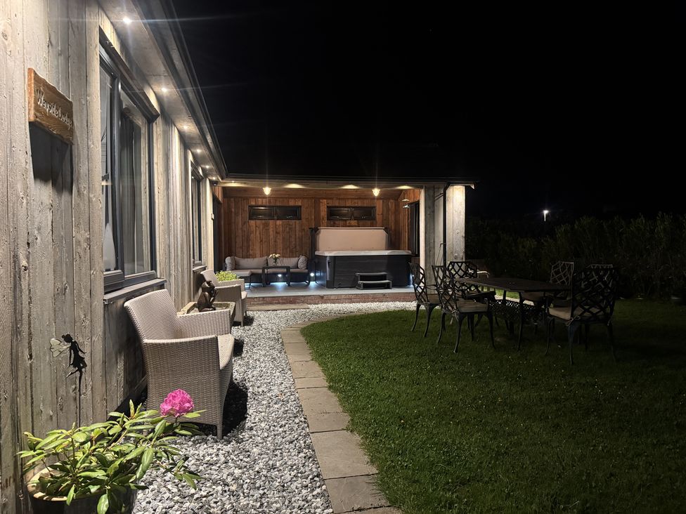 An outdoor patio at night with chairs on a gravel path a table with chairs on grass and a covered seating area with a hot tub at Wayside Lodge in Blackpool