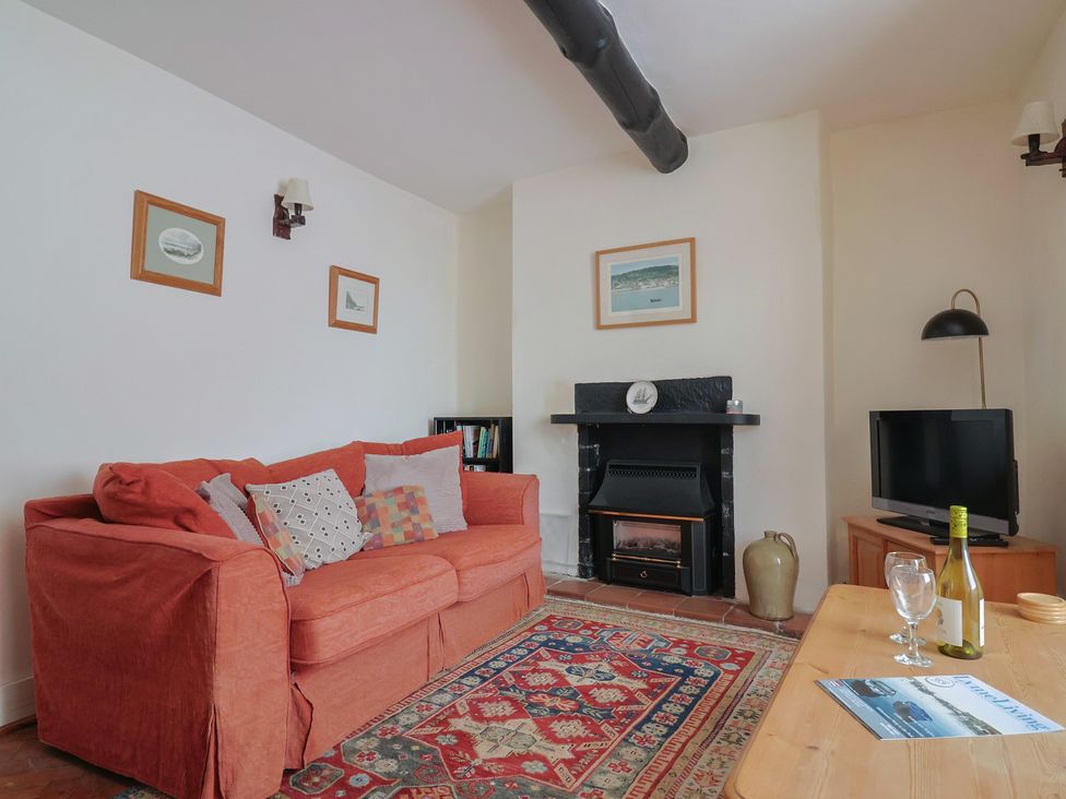 A living room with an orange sofa a patterned rug a fireplace a wooden table with wine bottle glasses and a magazine at Milton Cottage Lyme Regis