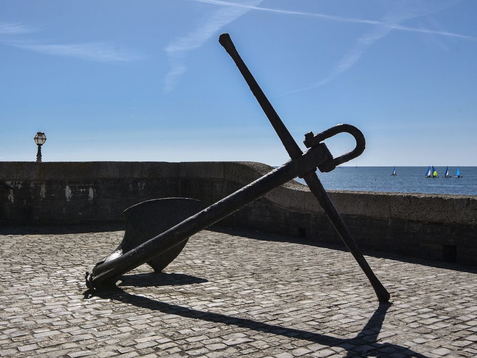 A large metal anchor on a paved platform by a stone wall with the sea and sailboats in the background at Milton Cottage in Lyme Regis