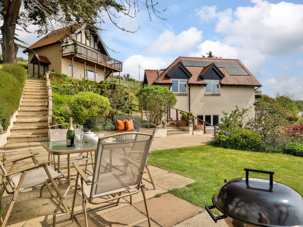 A garden patio with a table and chairs and barbecue near two houses with stairs and greenery at Beer View Cottage in Beer