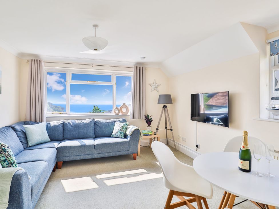 A living room with a blue corner sofa a round white table with a bottle and glasses a wall mounted tv and a window with a sea view at Beer View Cottage in Beer