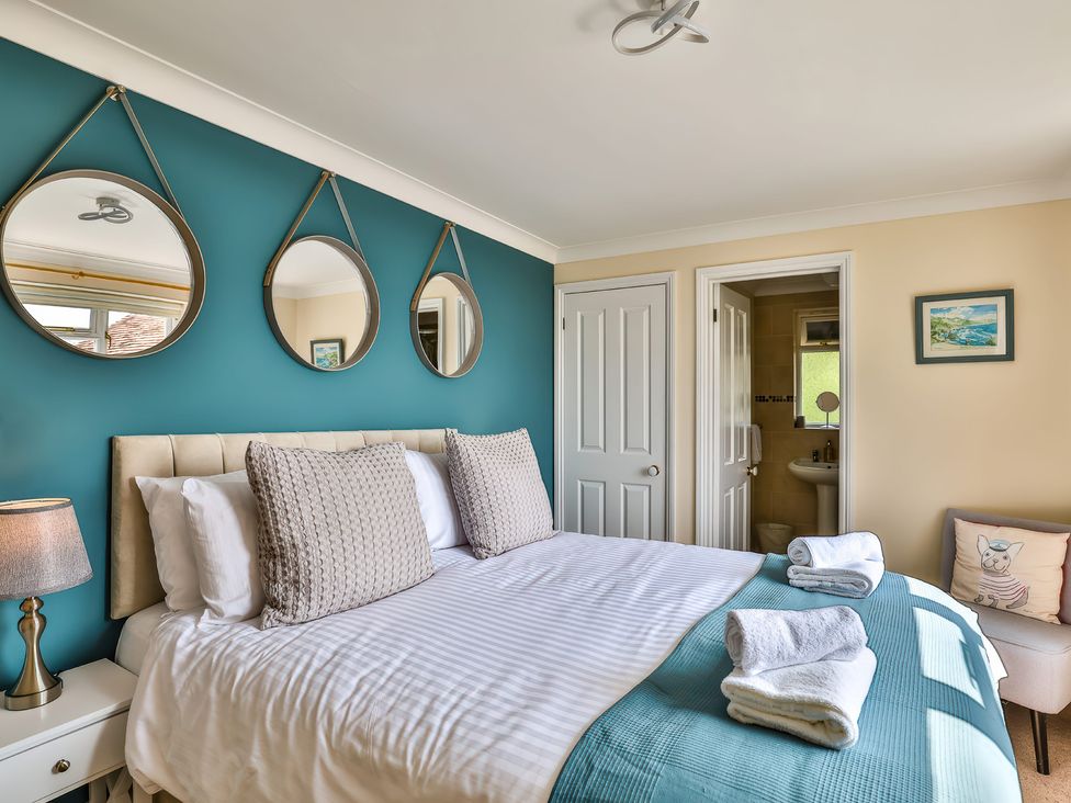A bedroom with a bed with pillows and folded towels a bedside table with lamp three round mirrors on a teal wall a door to a bathroom and a chair with a dog pillow at Beer View Cottage in Beer
