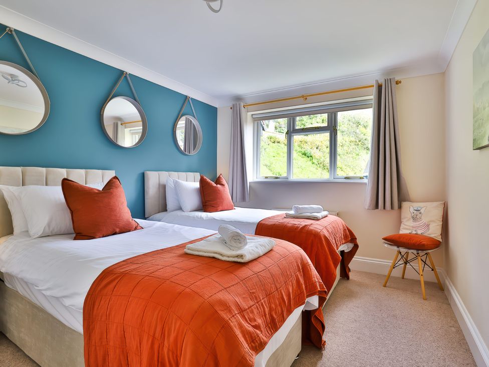 A bedroom with two single beds with orange blankets and pillows a blue accent wall with three round mirrors and a window with curtains at Beer View Cottage in Beer