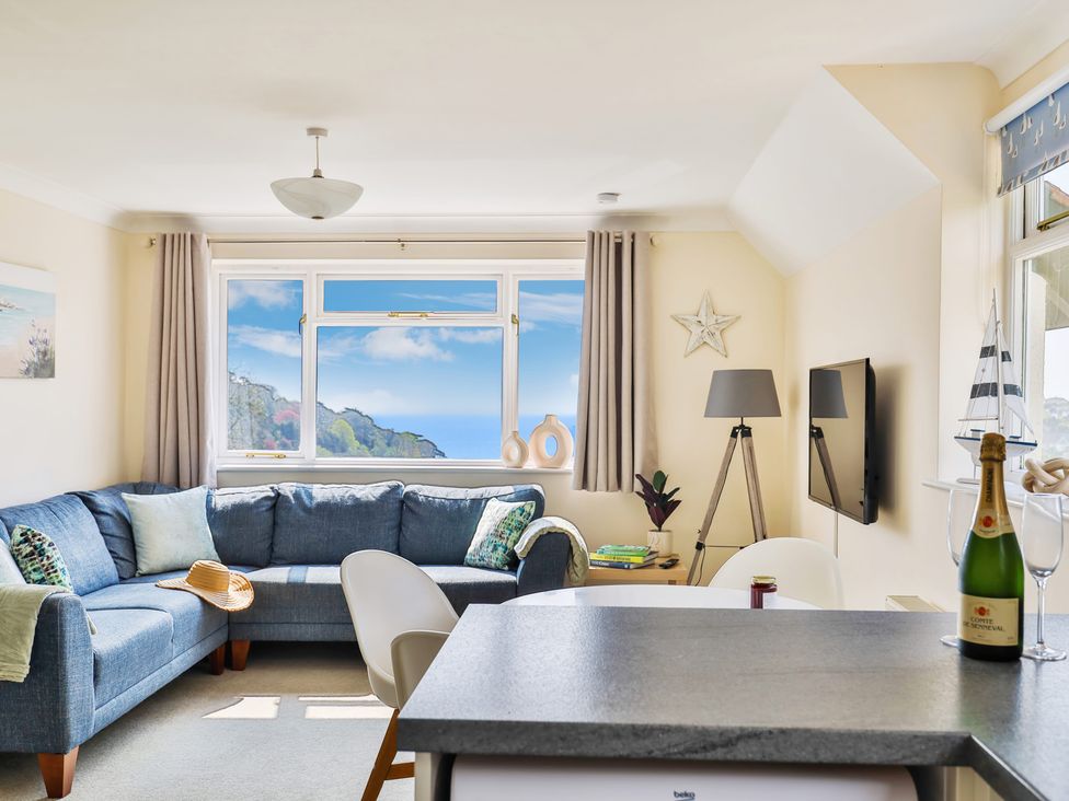 A living room with a blue corner sofa a window showing a sea view a floor lamp a wall-mounted TV and a kitchen counter with a bottle of champagne at Beer View Cottage in Beer