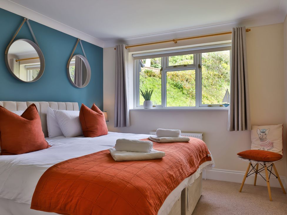 A bedroom with a double bed with orange pillows and blanket two round mirrors above the bed a window with curtains and a chair with cushions at Beer View Cottage in Beer