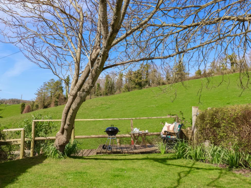 A seating area with a chair a small table and a barbecue grill on a wooden deck next to a tree and open green field at Beer View Cottage in Beer