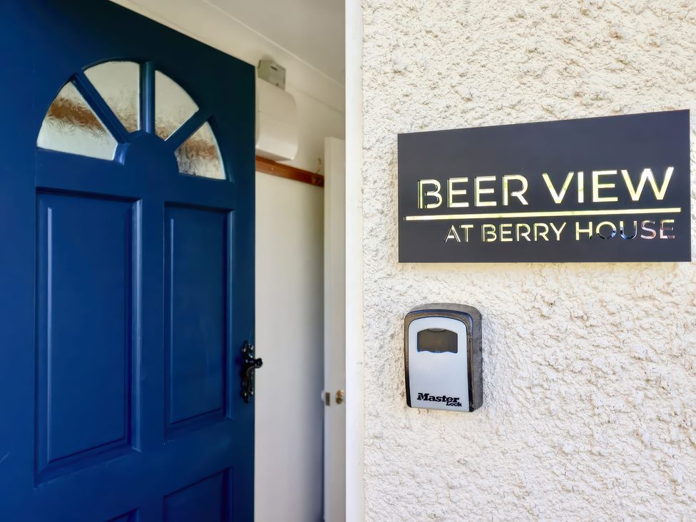 A blue door open next to a wall sign that reads Beer View at Berry House with a lockbox below the sign at Beer View Cottage in Beer