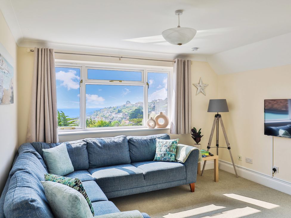 A living room with a blue sectional sofa a floor lamp a small wooden table and a wall-mounted TV at Beer View Cottage in Beer