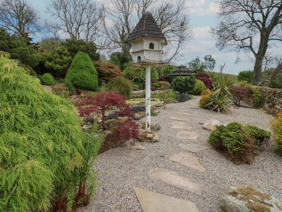 A garden with a gravel path and a birdhouse on a pole surrounded by bushes and trees at Pagecroft Cottage in Haydon Bridge