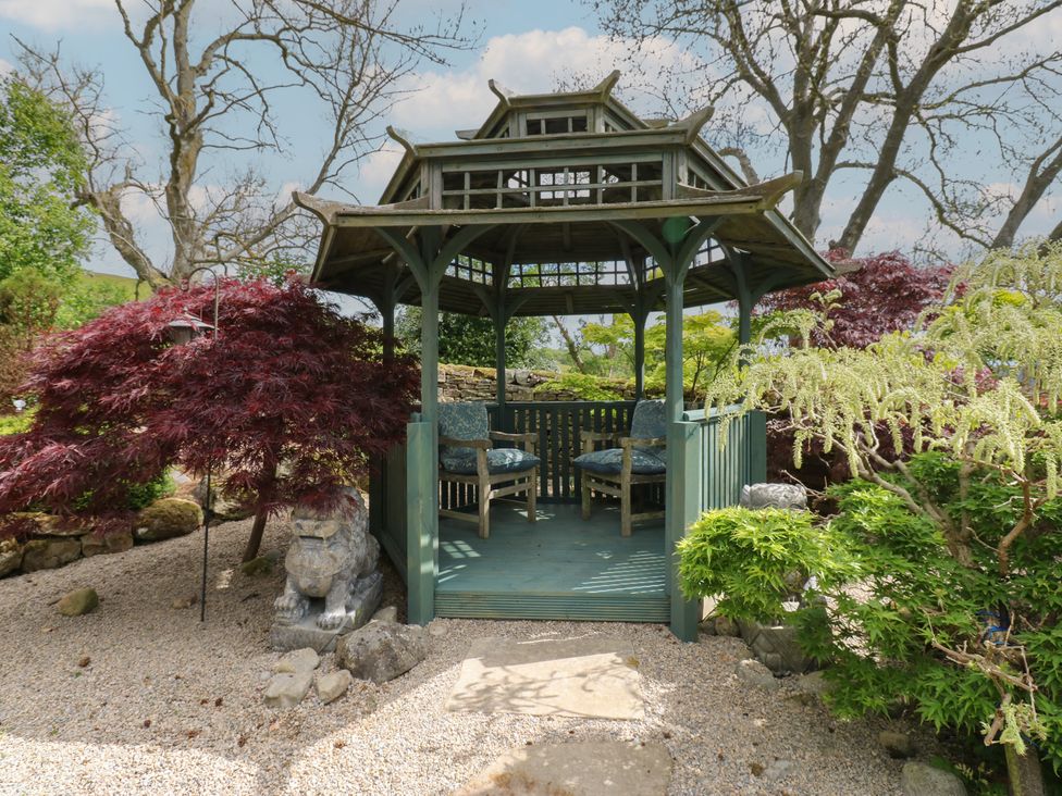 A wooden gazebo with two chairs inside surrounded by trees and shrubs at Pagecroft Cottage in Haydon Bridge