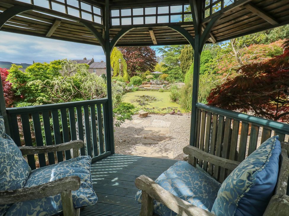 A garden view from a wooden gazebo with cushioned chairs overlooking a pond and plants at Pagecroft Cottage in Haydon Bridge