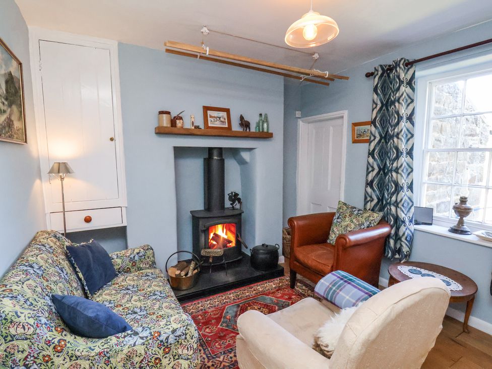 A small living room with a wood burning stove a patterned sofa two armchairs and a window with curtains at Whinstone Cottage Esk Valley near Grosmont