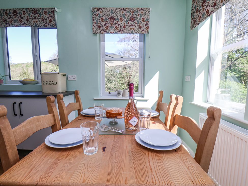 A dining area with a wooden table set with plates glasses a bottle of prosecco and a cake near windows with floral blinds at Whinstone Cottage Esk Valley near Grosmont
